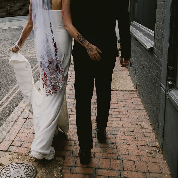Tattooed bride in embroidered veil walks a brick pavement beside a suited groom