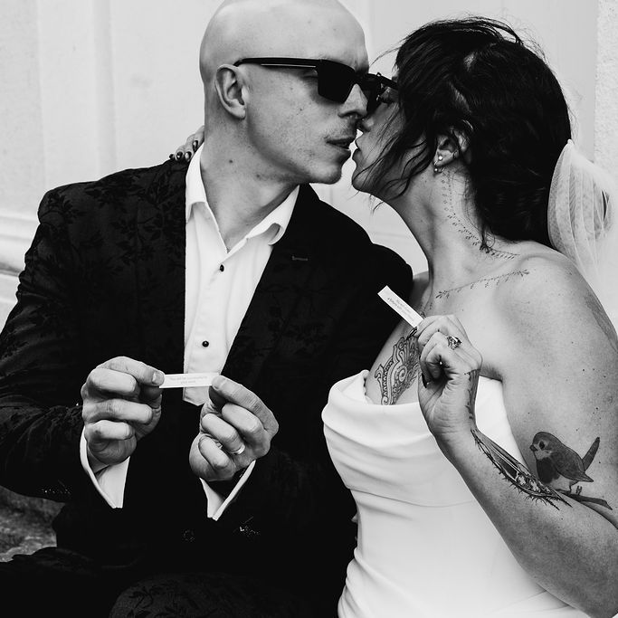 Bride and groom lean in to kiss while holding paper notes outside a period building