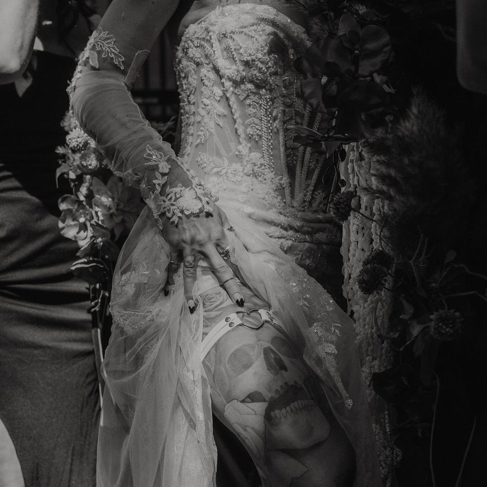 Tattooed bride lifts embellished lace gown to reveal skull leg tattoos in black and white