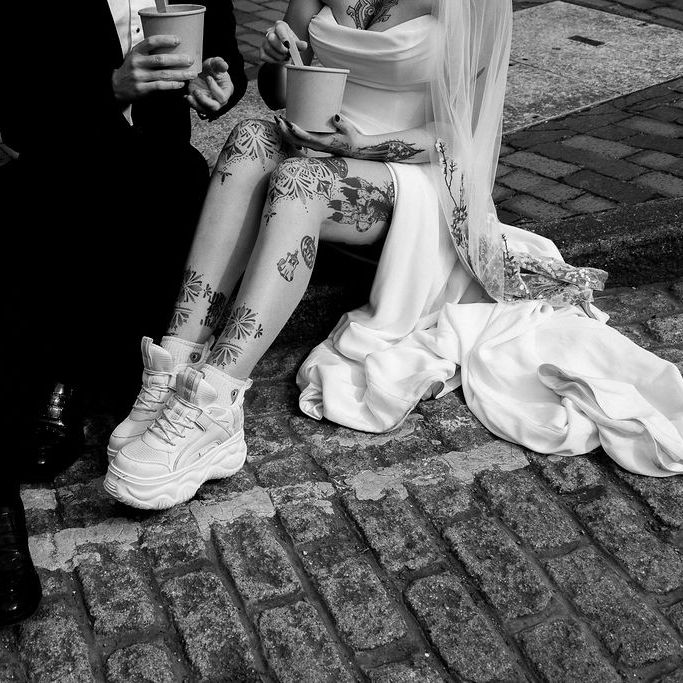 Tattooed bride in chunky trainers and groom sitting on kerb with drinks on cobbled street