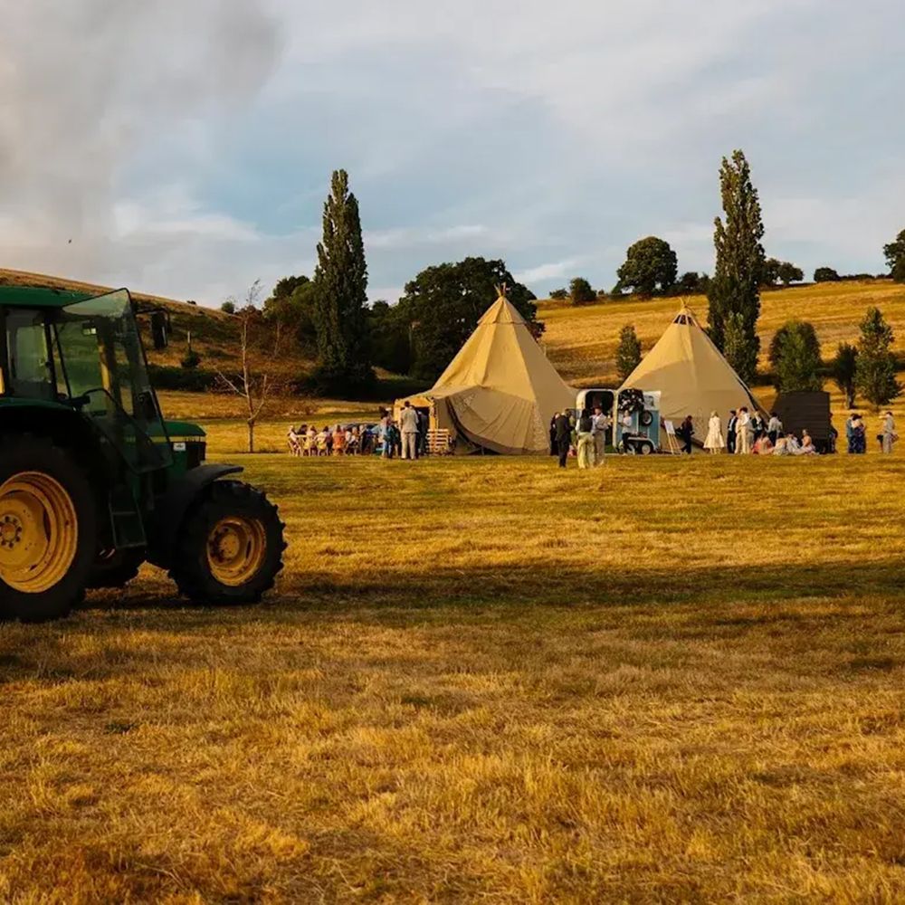 Green tractor and trailer in a golden field beside tipis where wedding guests gather at dusk