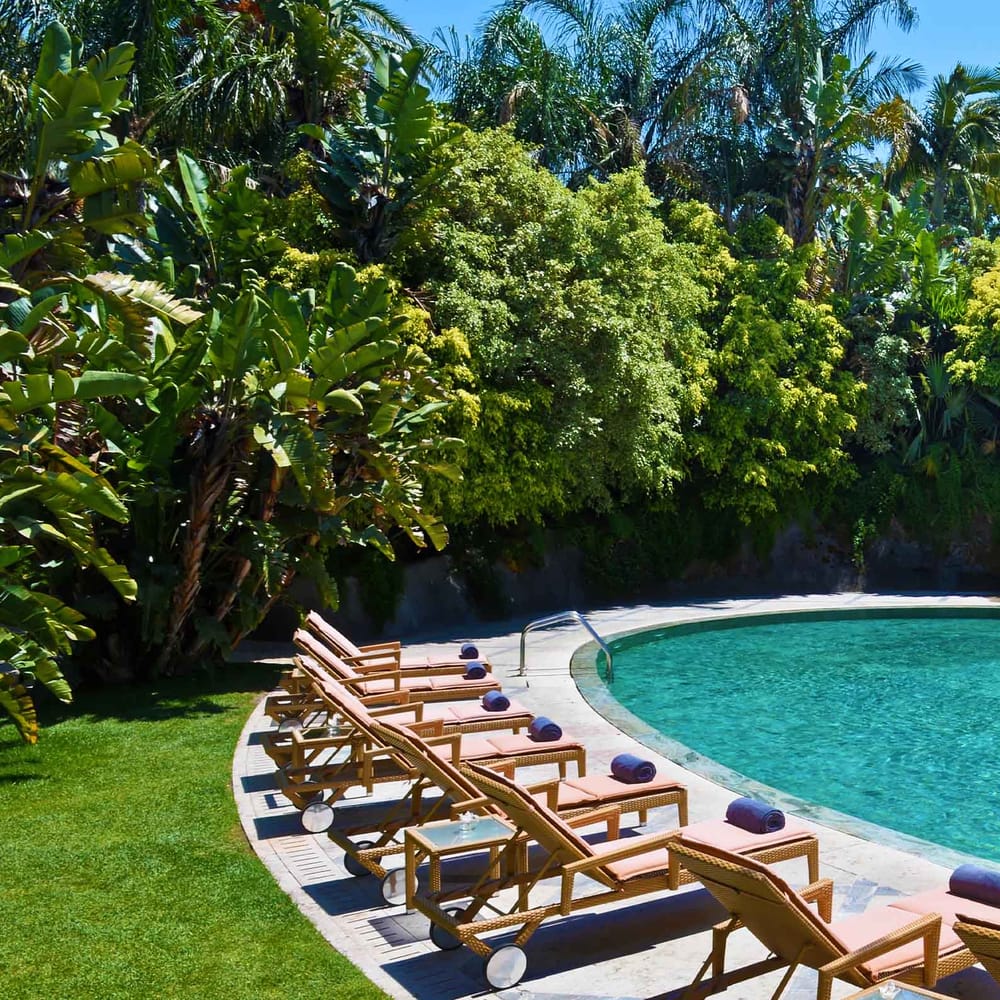 Curved outdoor pool with fountain, sun loungers and parasols beside a lush tropical garden and villa