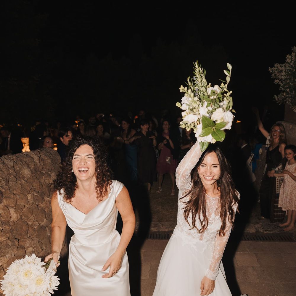 Two brides laughing and holding bouquets aloft as guests celebrate behind them at night