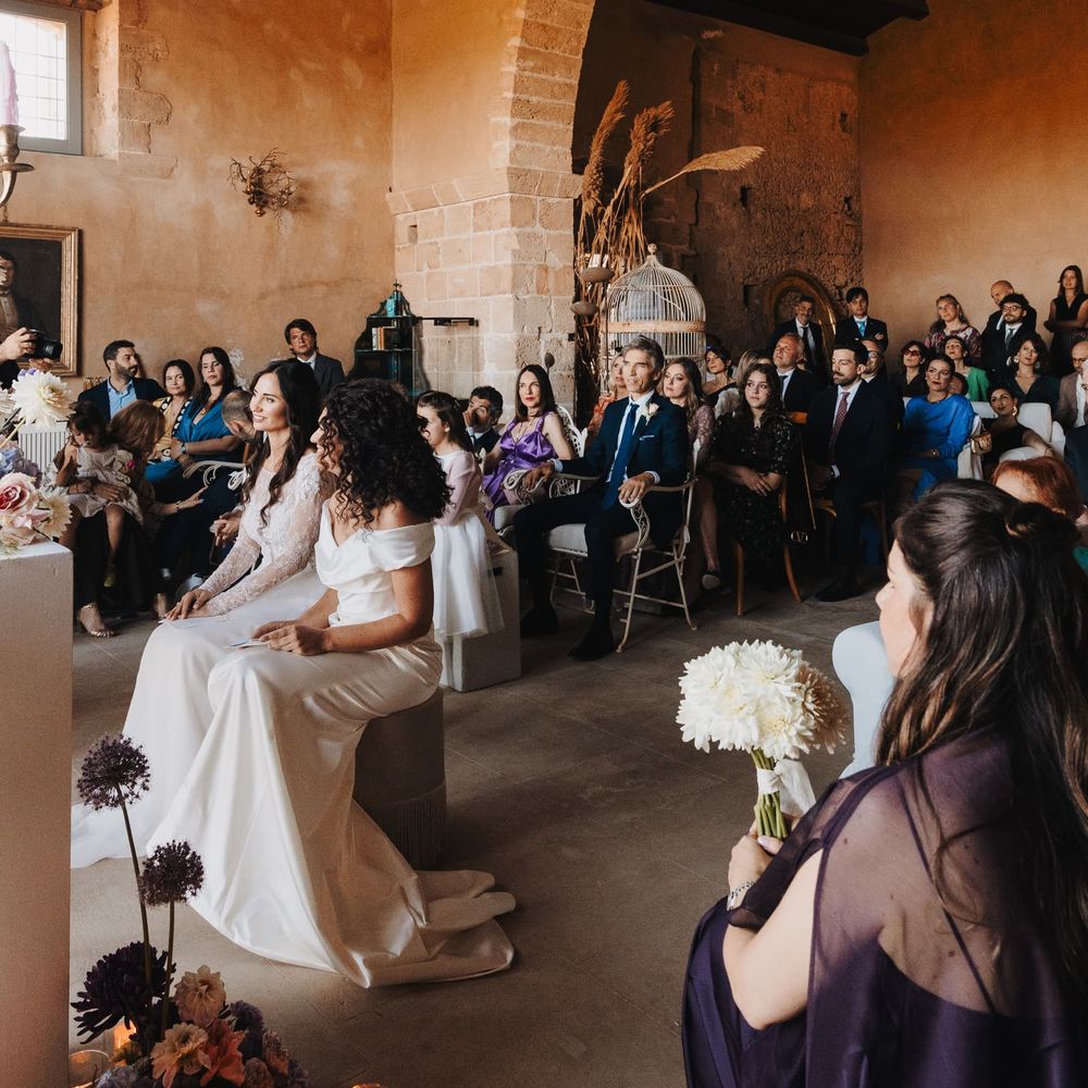 Two brides seated during a ceremony inside a rustic stone venue with a sweeping brick arch