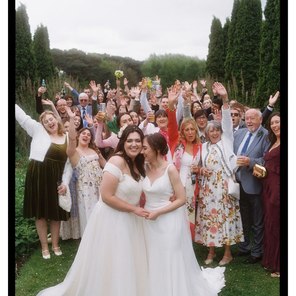 Two brides in white gowns stand together as guests cheer with raised hands in a garden