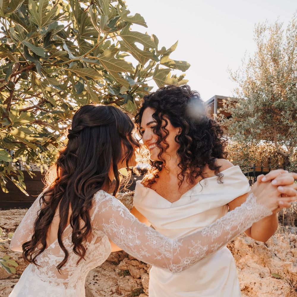 Two brides in ivory gowns hold hands and share a tender moment among olive trees at golden hour