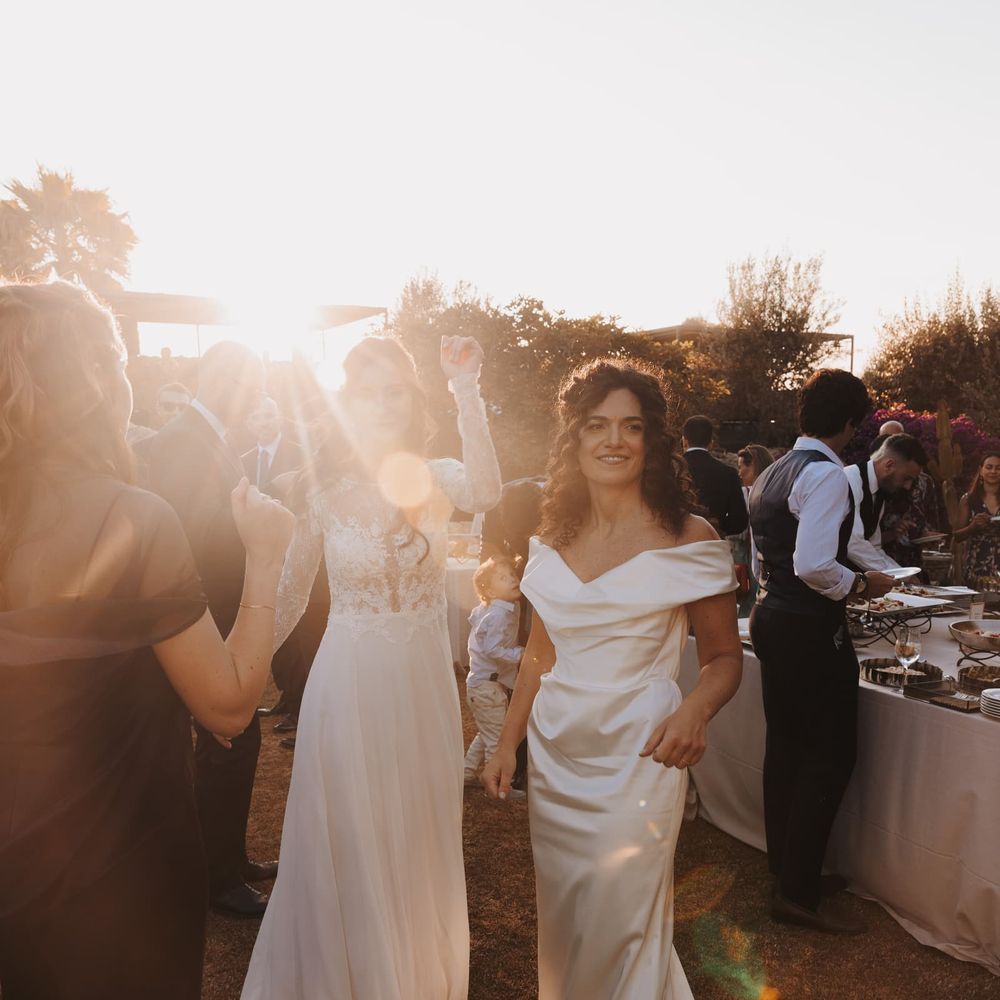 Two brides in white gowns dancing among guests at a golden-hour outdoor reception