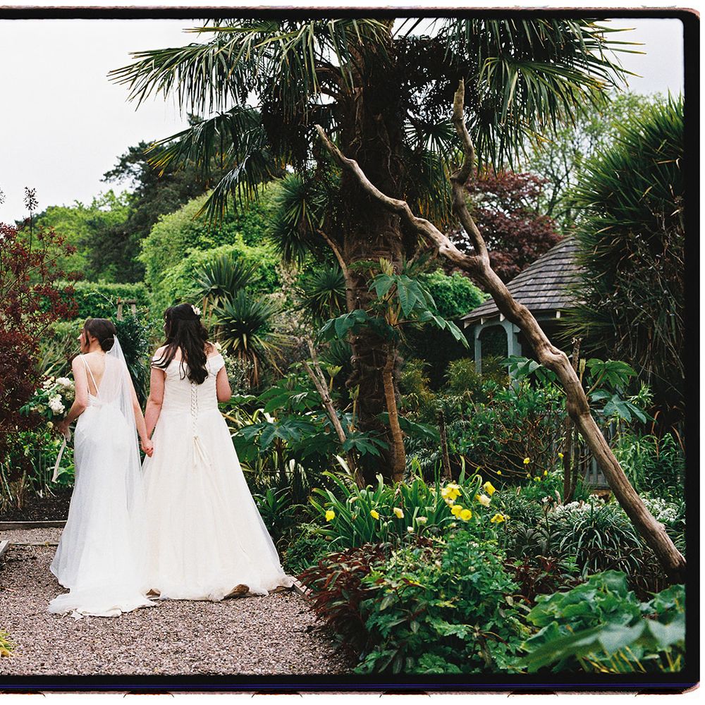 Two brides in white gowns holding hands on a gravel path through a lush subtropical garden