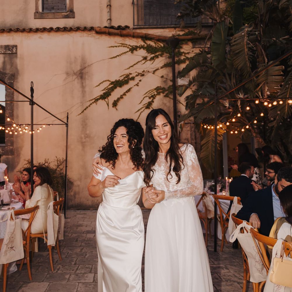 Two brides in white gowns laugh together in a courtyard reception at dusk