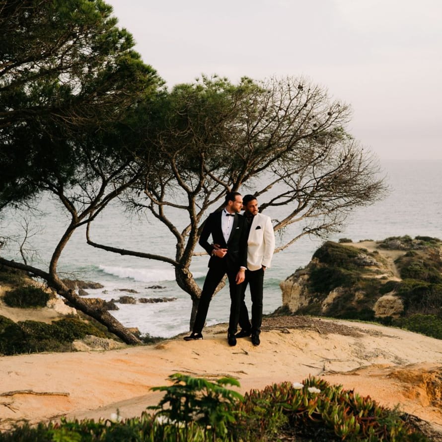 Two grooms in black and white tuxedos stand beneath a windswept pine on a coastal clifftop
