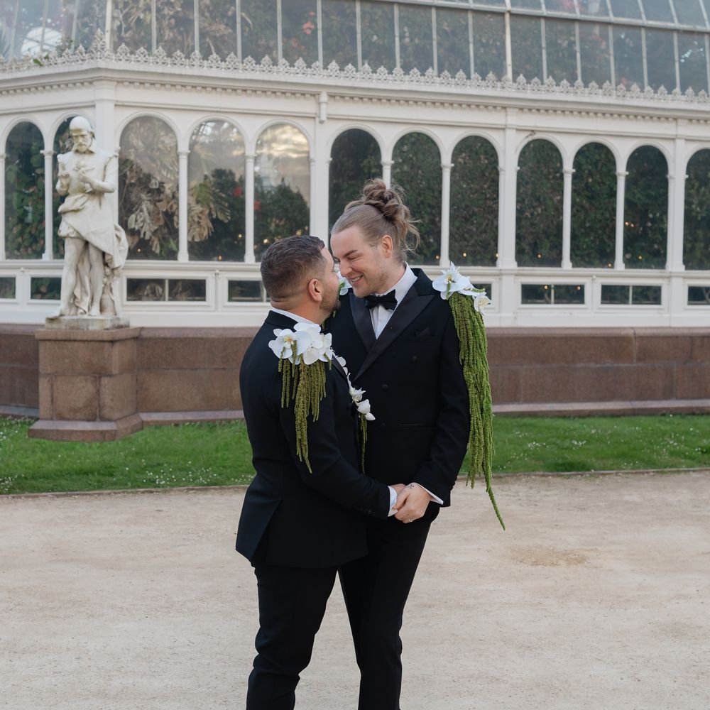 Two grooms in black tuxedos share a tender moment outside an ornate glasshouse conservatory.