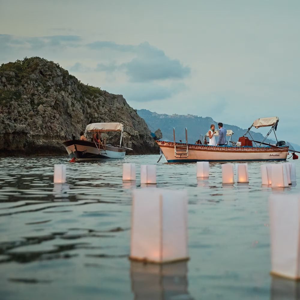 Couple aboard a wooden boat at dusk surrounded by floating paper lanterns on calm coastal water