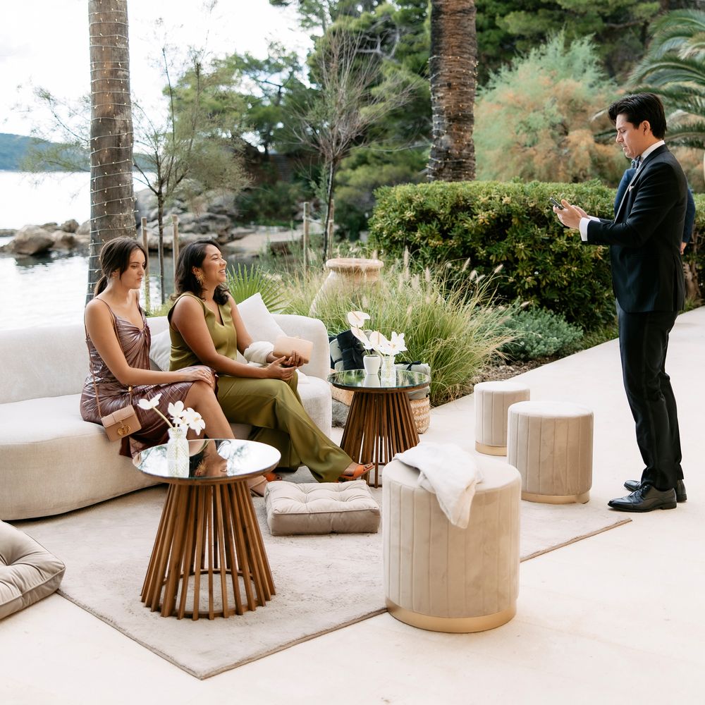 Two guests seated on a cream sofa in an outdoor lounge as a man in a suit stands nearby