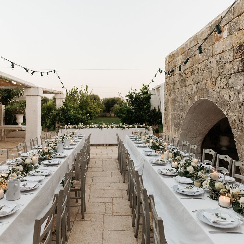Outdoor courtyard wedding breakfast with long tables, white florals and festoon lighting at dusk
