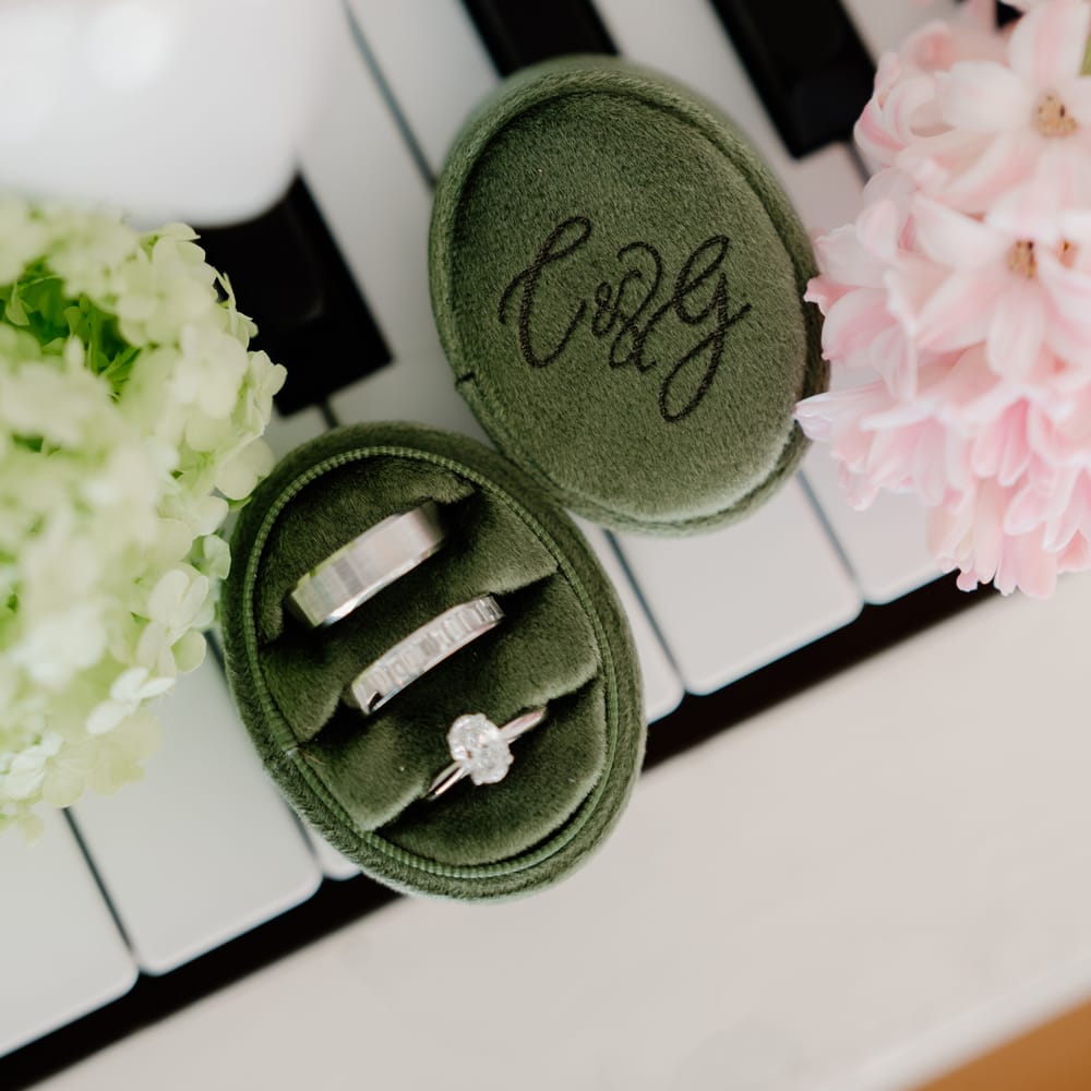 Wedding rings in a green velvet monogrammed box resting on piano keys with pink and green blooms