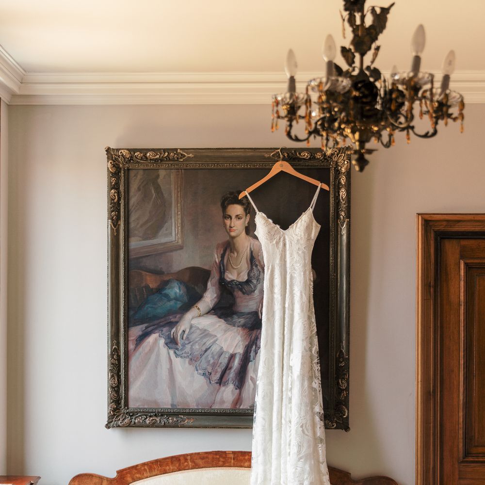 Lace wedding gown hanging before a framed portrait above an antique cream chaise longue