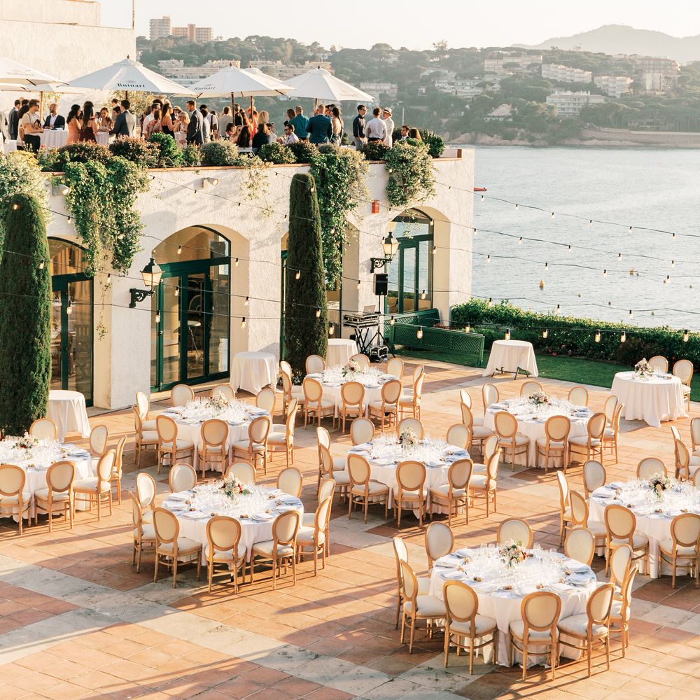 Coastal terrace set with round tables and gold chairs as guests gather under parasols at golden hour