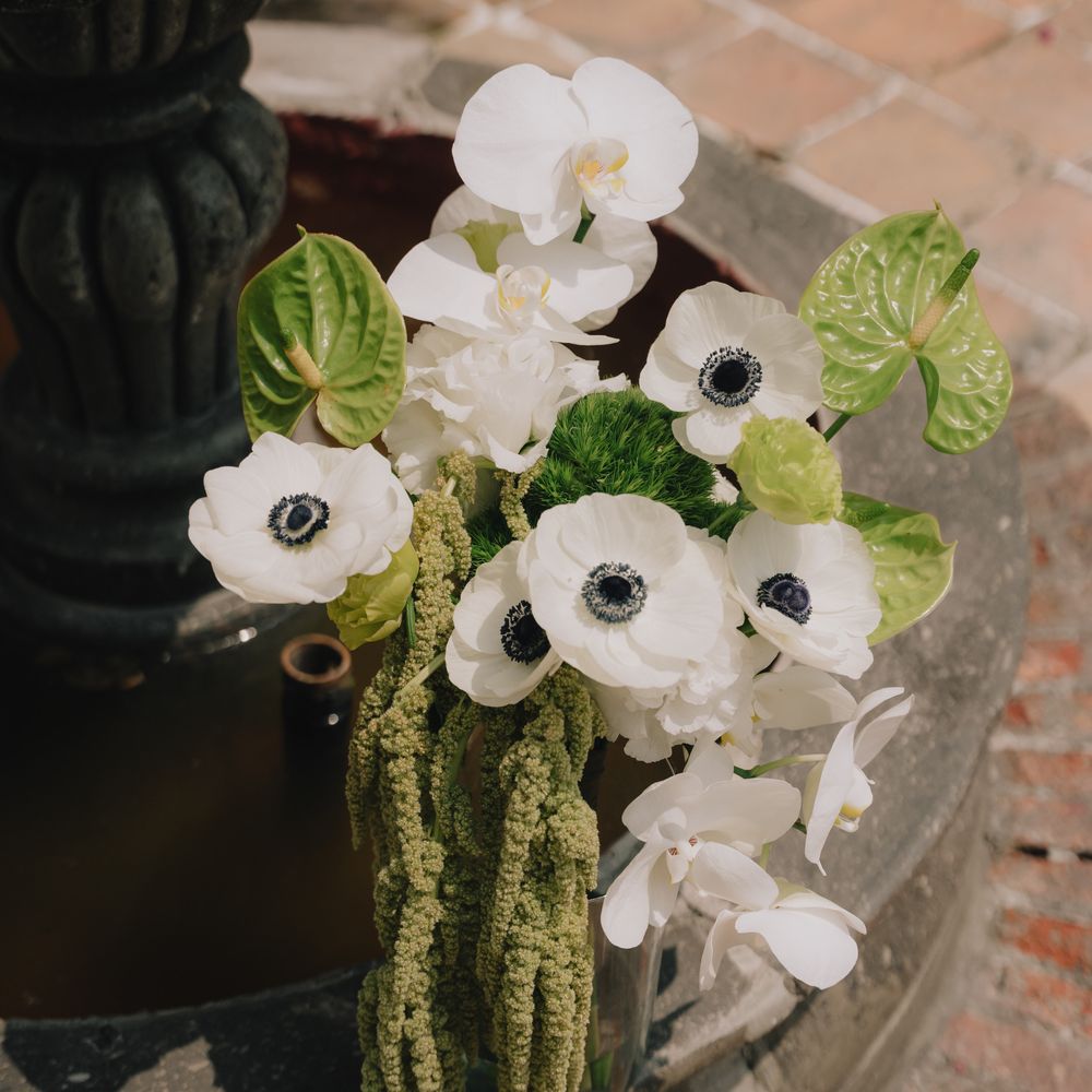 White anemones, orchids and green anthuriums with trailing amaranthus beside a stone fountain