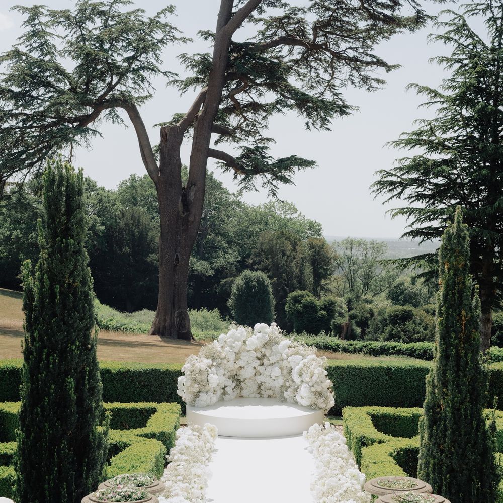 Outdoor ceremony aisle lined with white blooms leading to a floral arch in formal gardens