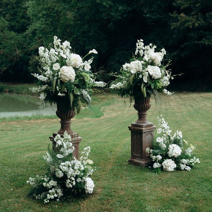 White floral arrangements in stone urns on plinths beside a lake on a green lawn