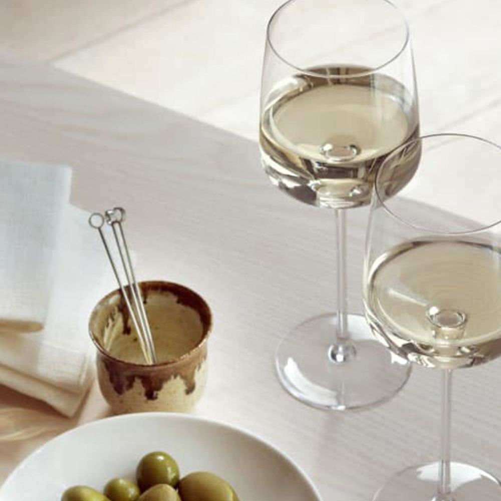 Two glasses of white wine beside a white bowl of green olives on a pale wooden table