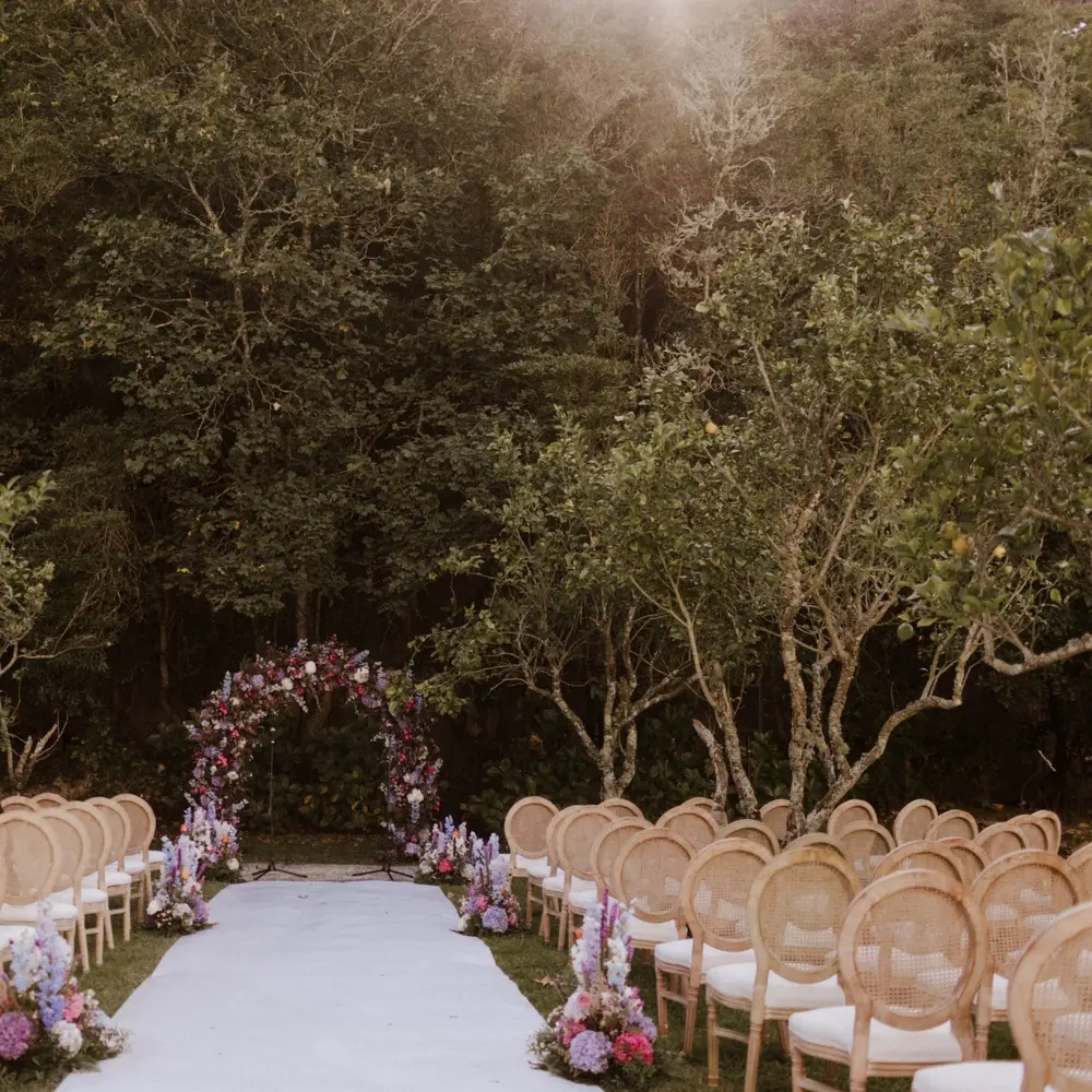 Floral arch and aisle flanked by rattan chairs set for an outdoor woodland ceremony