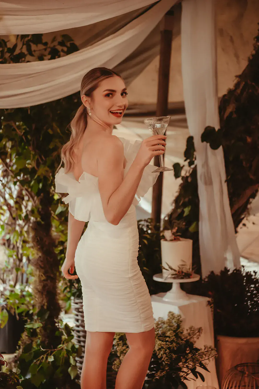 Bride in short wedding reception dress with frill detail sipping wedding cocktail with sleek ponytail 