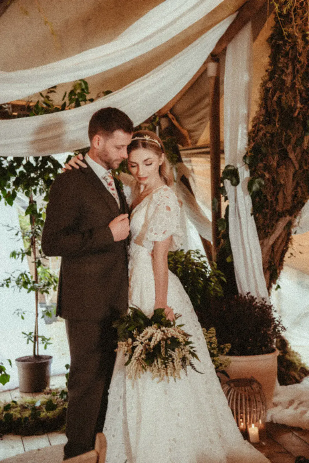 Groom in brown wedding suit with floral tie embracing the bride in a floral lace wedding dress and headband with foliage wedding bouquet