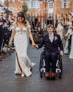 Bride in fitted wedding dress has confetti exit with groom in a blue suit in a wheelchair.