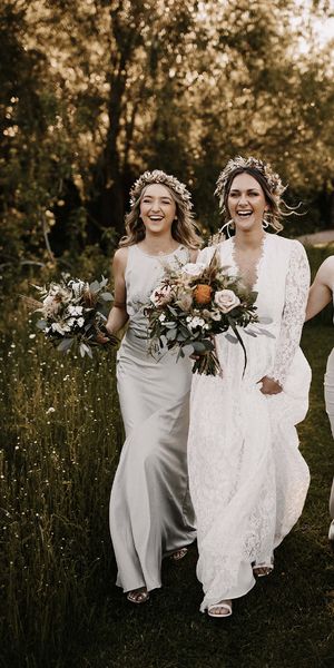 Bridesmaids in silver satin dresses and bride in a lace dress walking through the fields with a dried flower crown on their heads