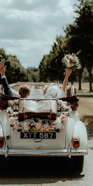 Bride and groom ride away in their wedding car with an old fashioned suitcase and flower decor.
