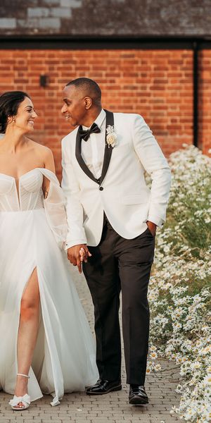 bride in off-the-shoulder tulle wedding dress and groom in black and white tuxedo walk hand in hand through white flowers at Crumplebury