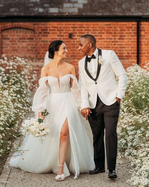 bride in off the shoulder tulle wedding dress and groom in black and white tuxedo walk hand in hand through white flowers at Crumplebury
