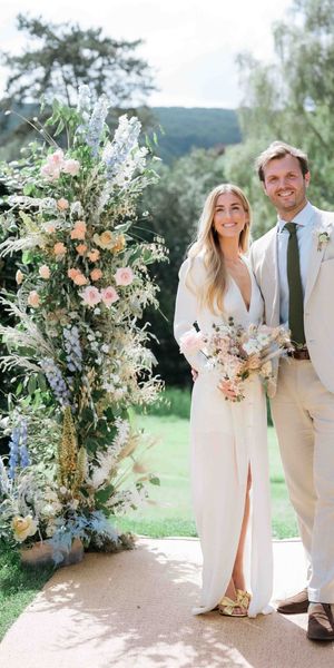Bride wearing custom Bon Bride gown with plunging neckline and front slit with groom in cream suit in front of pastel flower displays for garden tipi wedding 
