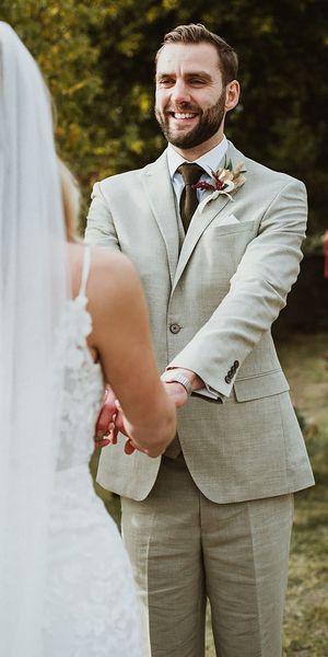 Groom wears classic stone beige linen suit, white shirt and wool green tie alongside a floral buttonhole