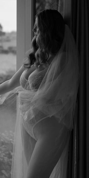 Bridal boudoir portrait of a bride wearing delicate ivory lace lingerie and a cathedral-length veil, standing beside a floor-to-ceiling window overlooking the Hampshire countryside. Captured in soft natural light with a fine art, black and white aesthetic. Photography by Rebecca Frost Photography.