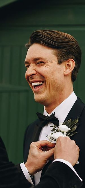 groom and his best man wearing black tie wedding suits while best man helps groom with buttonhole