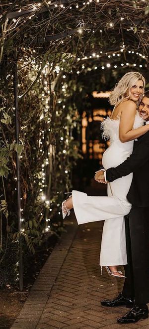 bride being picked up by groom wearing white bridal jumpsuit with feather trim