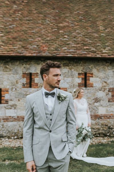 Portrait of the groom in a grey three-piece wedding suit with his bride in the background