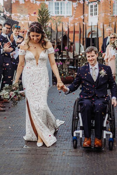 Bride in fitted wedding dress has confetti exit with groom in a blue suit in a wheelchair.