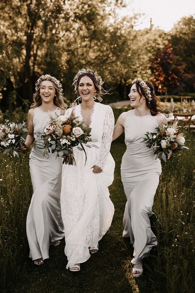 Bridesmaids in silver satin dresses and bride in a lace dress walking through the fields with a dried flower crown on their heads