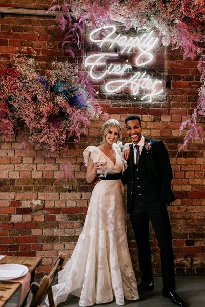 Prezola wedding couple standing in front of a neon happily ever after sign surround by pink gypsophila and wearing a ruffle sleeve wedding dress