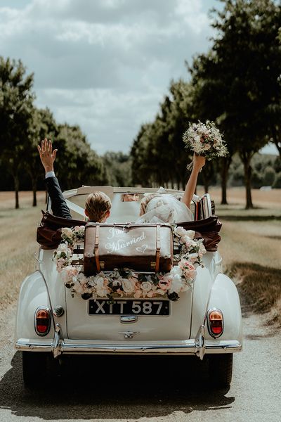 Bride and groom ride away in their wedding car with an old fashioned suitcase and flower decor.