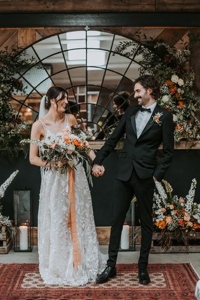 bride in a lace dress and groom in a black tuxedo at new Lake District wedding venue with oval micro back drop and colourful floral arrangements 