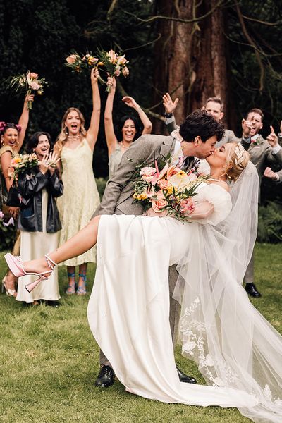 Groom dips bride in pink wedding shoes for a kiss with wedding party cheering in the background