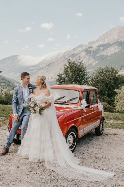 Bride and Groom lean against vintage red mini with the French Alps as their backdrop. Bride wears Halfpenny London gown.