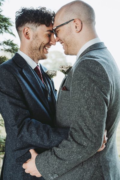 Grooms in blue and grey suit jackets with rose buttonholes made from pages from their favourite books for their DIY wood slice table centrepiece decor wedding.