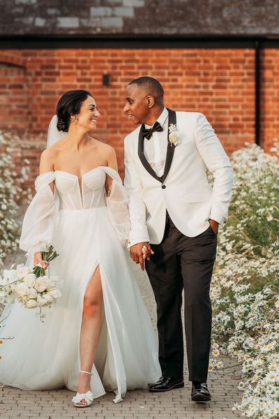 bride in off-the-shoulder tulle wedding dress and groom in black and white tuxedo walk hand in hand through white flowers at Crumplebury