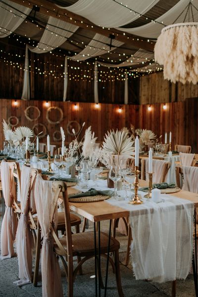 Romantic East Yorkshire Barns wedding reception with pink chiffon chair back decor, pampas grass chandelier, fairy light backdrop and dried palm leaf centrepieces 