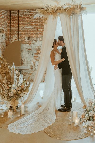 Bride and groom share a kiss under their wedding drapes at the altar decorated with candles at Godwick Barn. 